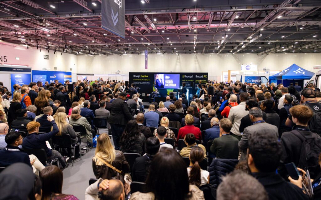 A standing audience facing the main stage at The Business Show London, with exhibition stands either side